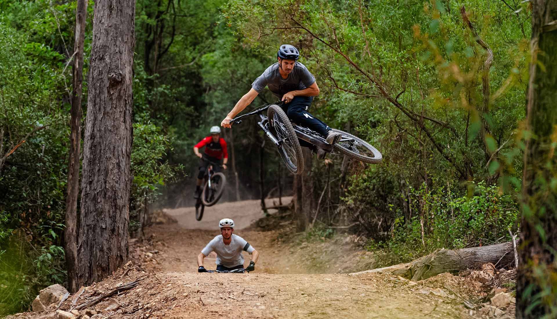 Three mountain bike riders jumping whoops on a forest trail